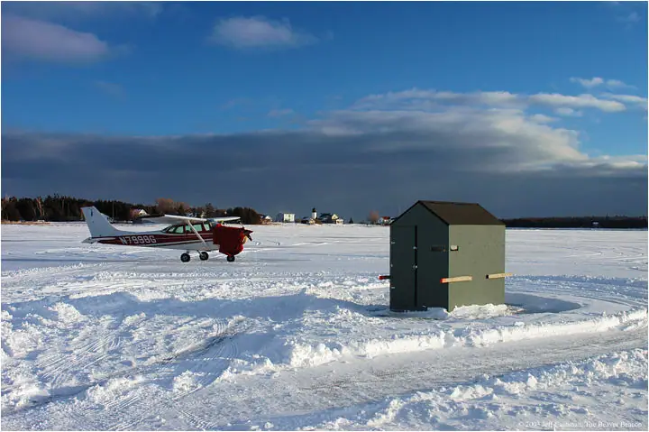 A small red-and-white airplane parked beside a dark green shed on a snow-covered open area.