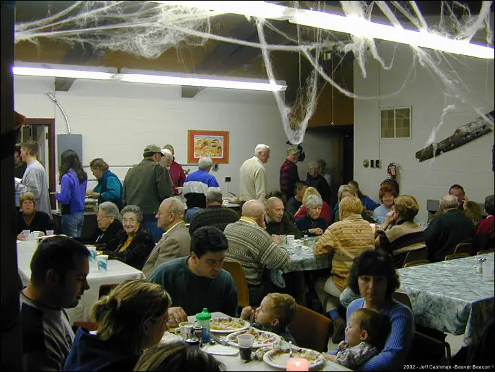 People gather around tables for a meal beneath hanging artificial cobweb decorations.