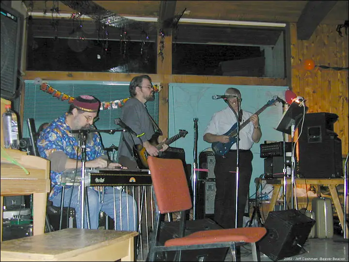 Three musicians perform indoors among instruments, amplifiers, chairs, tables, and string decorations.