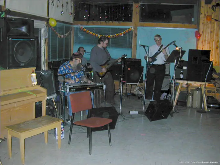 Four musicians perform indoors among instruments, speakers, balloons, and festive garlands.