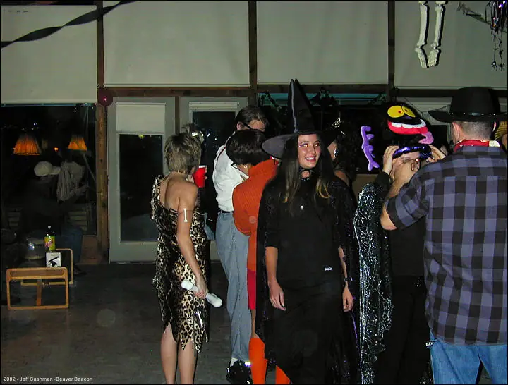 Adults in costumes gather indoors, including a woman wearing a pointed witch hat and black outfit.