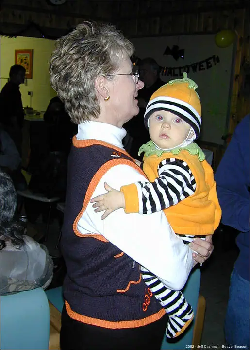 A woman holds a baby in a pumpkin costume at an indoor Halloween gathering.