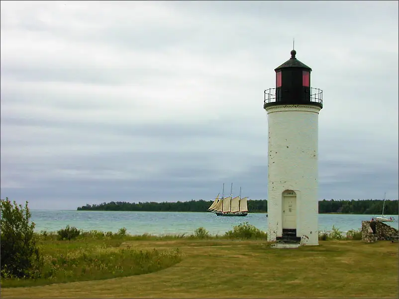 A white lighthouse stands beside the water as a multi-masted sailing vessel passes offshore.