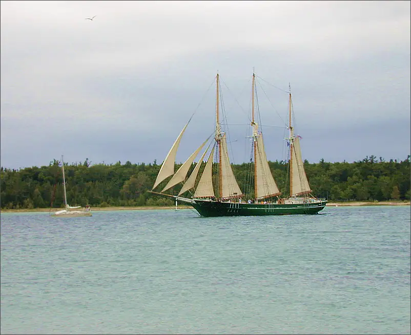 A three-masted sailing vessel with tan sails travels past a wooded shoreline beside a smaller sailboat.