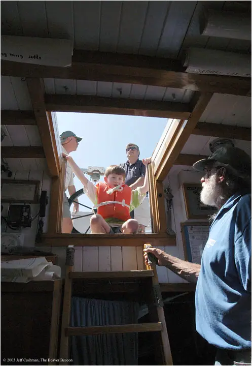 A child in an orange life vest sits in an open hatchway while adults look on inside a wooden vessel.