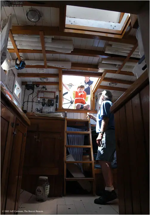 A child in an orange life vest and an adult stand inside a wood-paneled boat cabin beneath an open hatch.