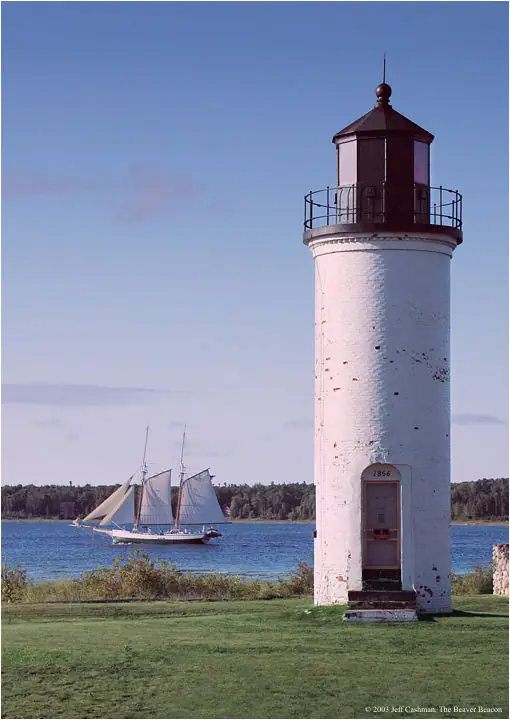 A white cylindrical lighthouse stands beside the water as a sailing vessel passes behind it.