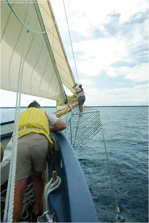 Two people aboard a sailboat beneath a large pale sail, with open water and a partly cloudy sky beyond.