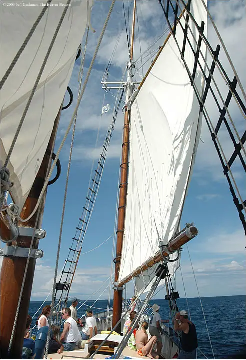 Tall wooden masts, white sails, rigging, and passengers fill a schooner viewed from the deck.