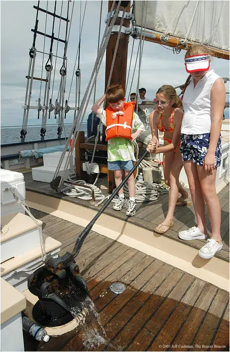 Children and adults operate a hand pump on the wooden deck of a sailing vessel.