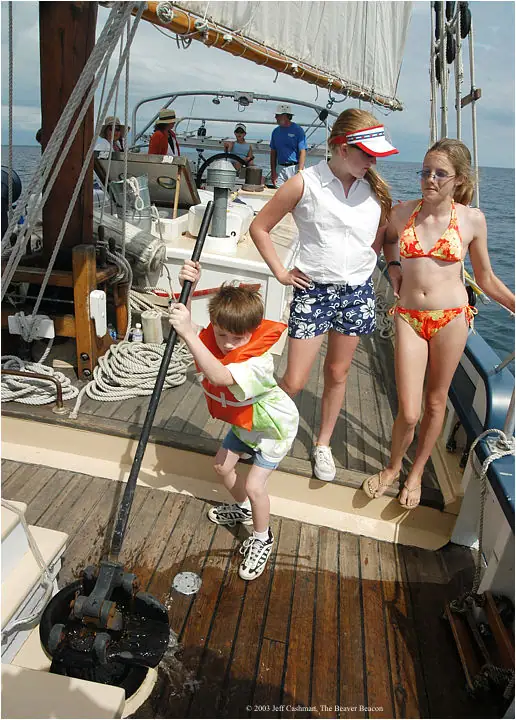 People gather around a child operating a pump on the wooden deck of a sailing vessel.