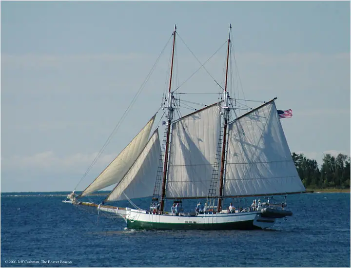 A two-masted schooner with white sails passes a wooded shoreline.