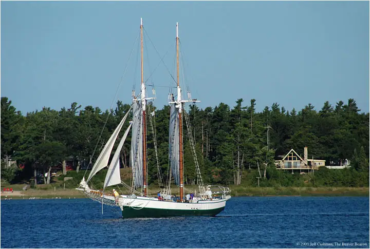 A two-masted schooner sails past a wooded shoreline with a house visible behind it.