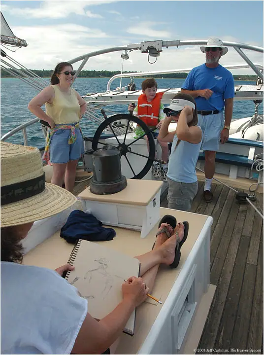 People gather on a sailboat deck while a seated person sketches in a spiral-bound notebook.