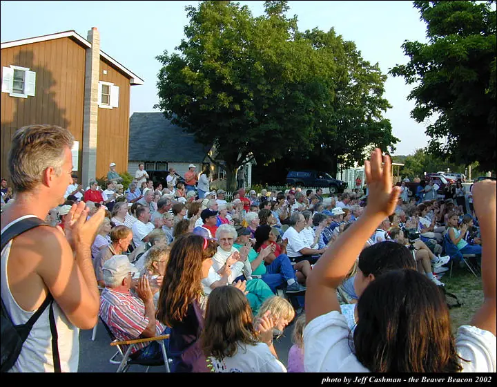 A large outdoor crowd sits in folding chairs, clapping and waving beside a wooden building.