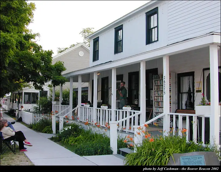 A person holds a microphone on the porch of a white two-story house while another person sits nearby.