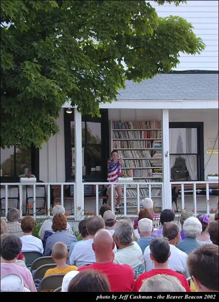 A woman in a flag-patterned shirt addresses a seated crowd from a porch.