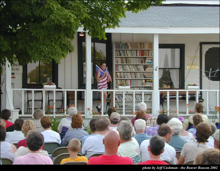 A person in a flag-themed shirt addresses a seated crowd from a porch lined with bookshelves.