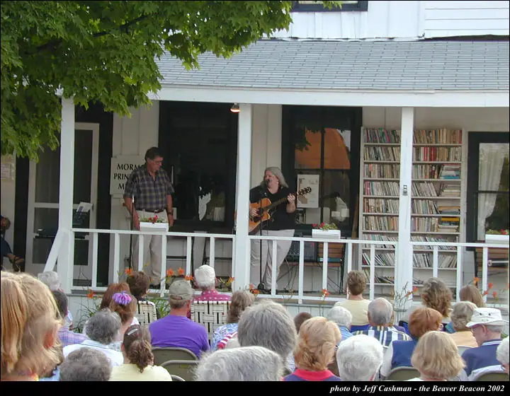 A guitarist performs on a porch before a seated audience while a man stands nearby.