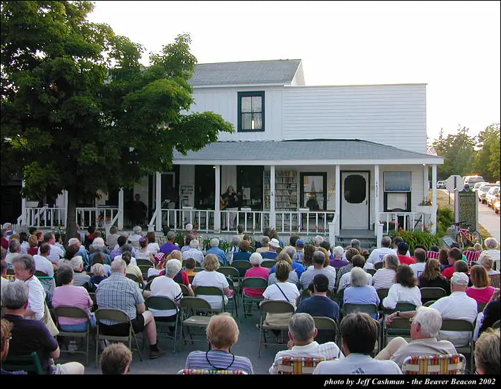 A large crowd watches a musician performing on the porch of a white building.