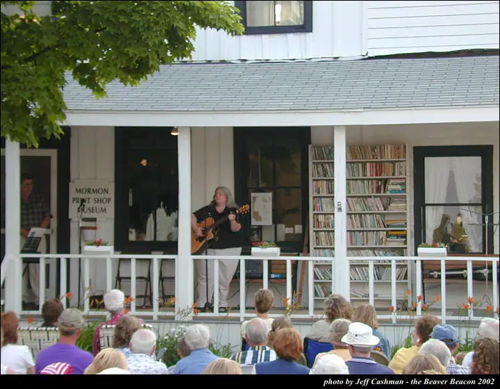 Musician playing guitar on a porch before a seated crowd.