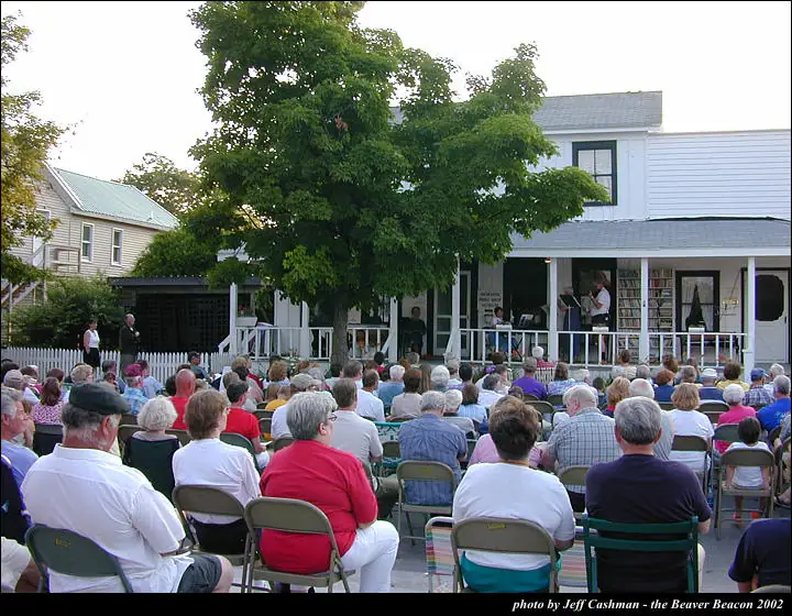 A large seated crowd watches performers on the porch of a house beneath a leafy tree.
