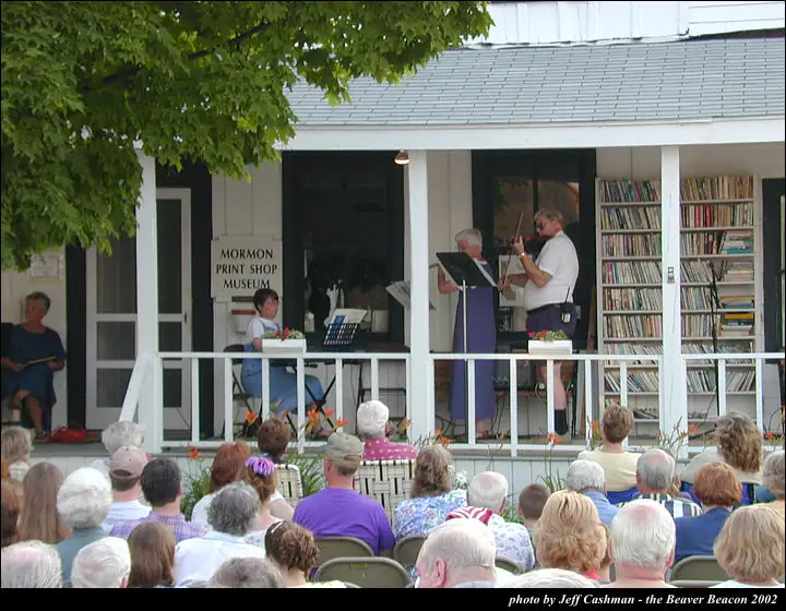 Two musicians perform on a porch before a seated audience.