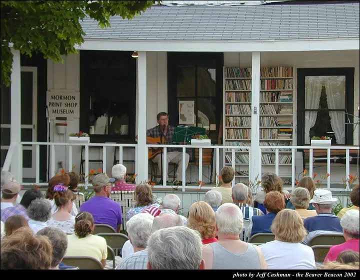 A guitarist performs on a porch before a seated outdoor audience.