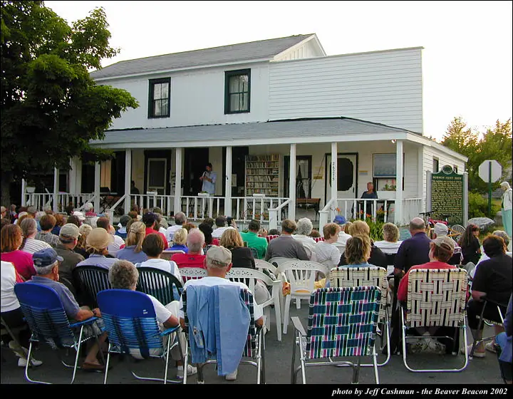 A large crowd sits in lawn chairs facing a person on the porch of a white building.