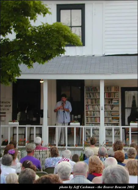A man speaks into a microphone from a porch while a crowd watches.