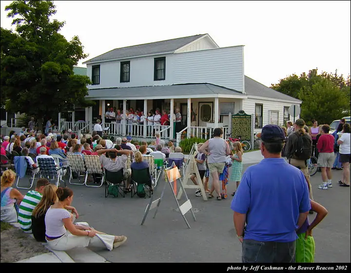 A large crowd gathers before a white-sided building where people stand on its covered porch.