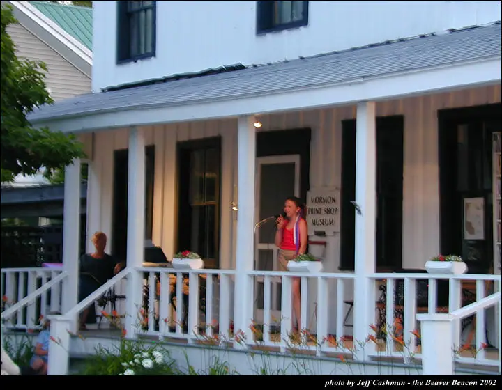 A woman sings into a microphone on a flower-decorated porch beside a seated person.