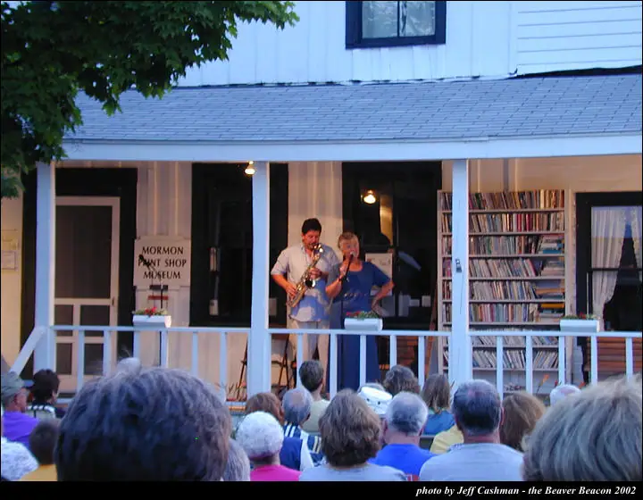 Two musicians perform on a porch before a seated audience.