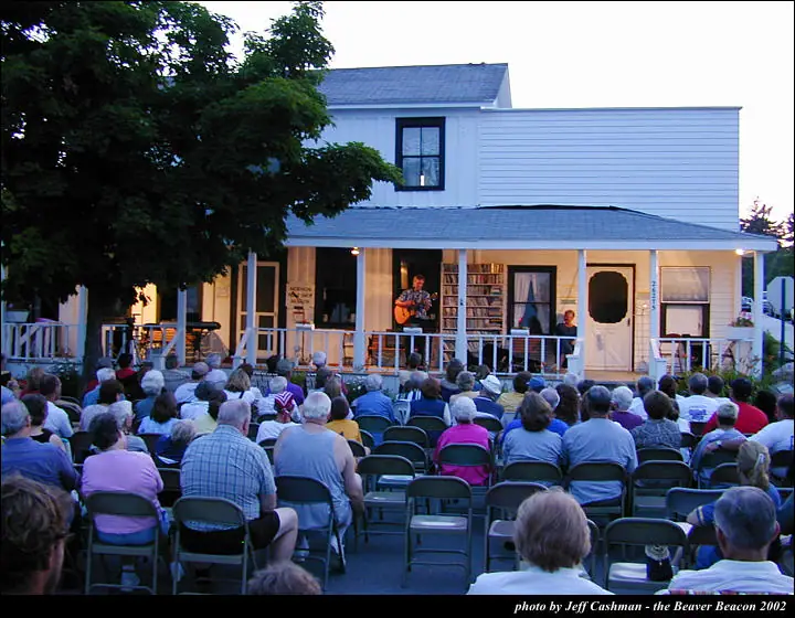 A guitarist performs on a house porch before a seated outdoor audience at dusk.