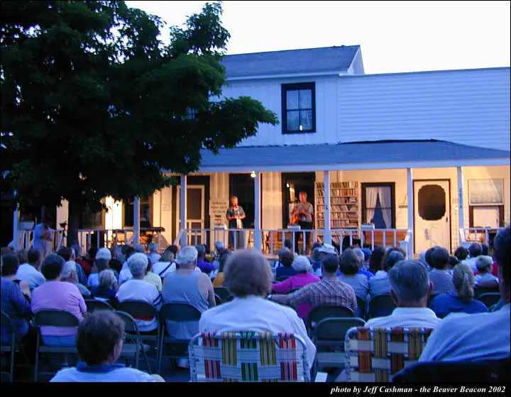 A seated crowd watches two musicians perform on the porch of a white building.