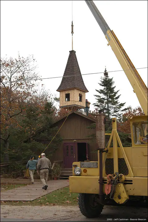 A crane lifts a wooden church steeple while workers stand beside the building.