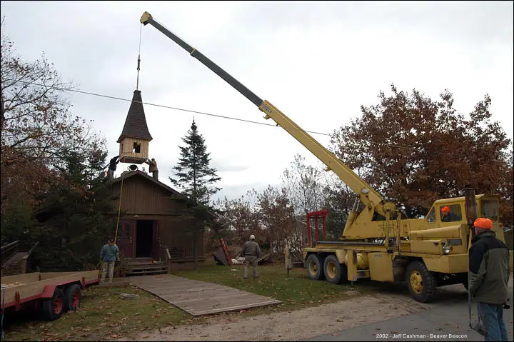 A crane lifts a steeple onto a wooden church while workers watch nearby.