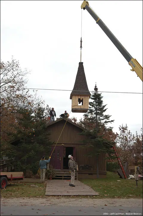 A crane suspends a wooden steeple above a small wooden church as people guide the installation.