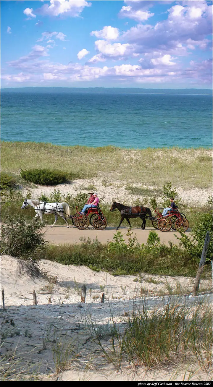 Two horse-drawn carriages travel along a sandy shoreline beneath a bright, cloud-filled sky.