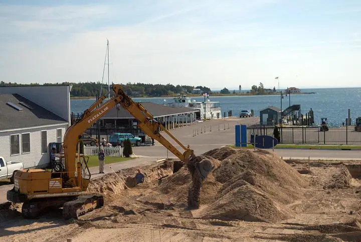 A tracked excavator moves sand at a waterfront construction site beside a dock and parked vehicles.