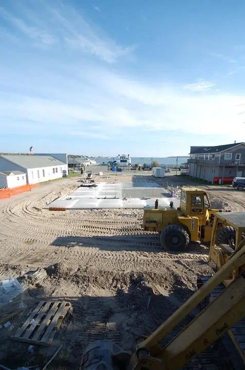 Construction equipment stands beside a rectangular concrete slab near waterfront buildings.