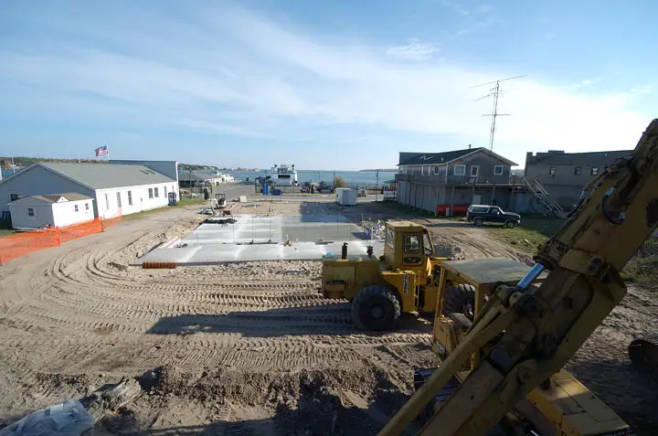 Construction equipment surrounds a freshly poured concrete slab beside waterfront buildings.