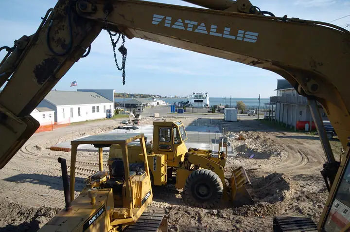 Construction equipment works on a dirt site beneath a large excavator arm, with buildings and waterfront visible beyond.