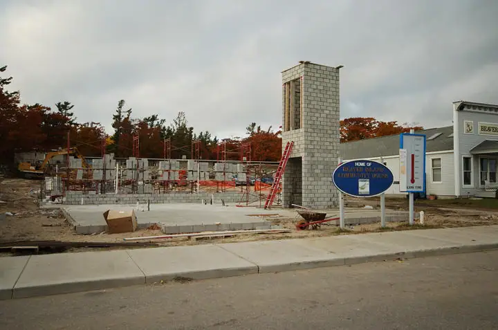 Concrete-block building under construction with scaffolding, ladders, a tall chimney, and a roadside sign.