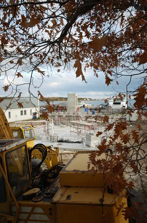 Construction site viewed through autumn branches, with a yellow excavator and concrete blockwork.
