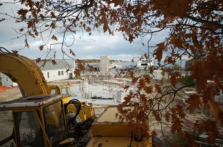 A yellow excavator overlooks a concrete-block building under construction, framed by autumn leaves.