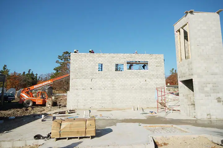 Concrete-block building under construction with an orange lift truck, scaffolding, and workers beneath a clear blue sky.