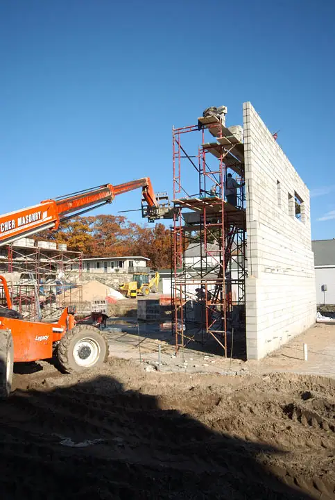 A masonry construction site with concrete-block walls, scaffolding, workers, and an orange lifting machine.