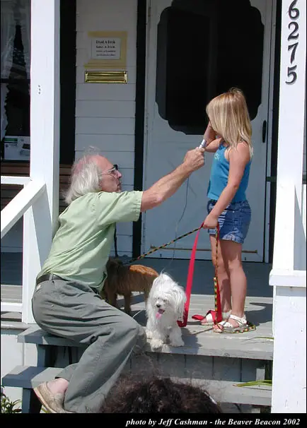 An adult kneels with two leashed dogs beside a child on a house porch.