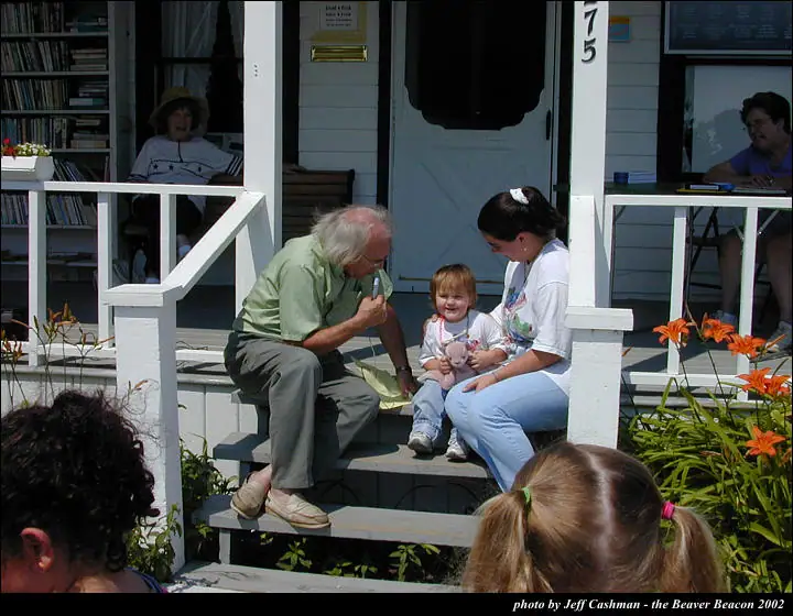 Adults and children gather on the steps of a white-painted building beside orange flowers.